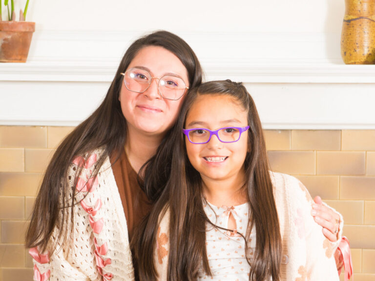 Mother with glasses hugging her daughter, wearing purple glasses, in front of a fireplace.